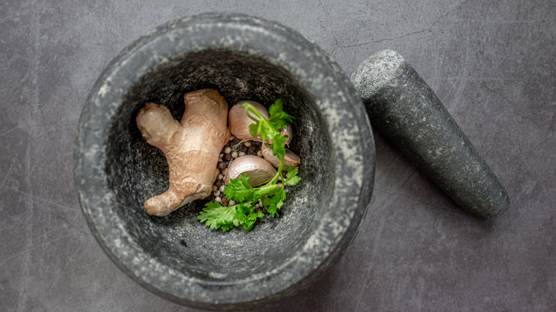 stone mortar containing spice paste ingredients (ginger, garlic, cilantro and spices), with its mortar sitting alongside on a dark stone countertop