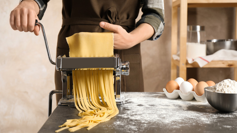 woman in a brown apron cutting fresh-made pasta into tagliatelle using a hand-cranked pasta roller, with eggs and flour on the counter beside her