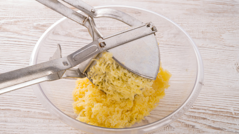a stainless steel potato ricer, shown in the midst of ricing cooked potatoes into a glass bowl.