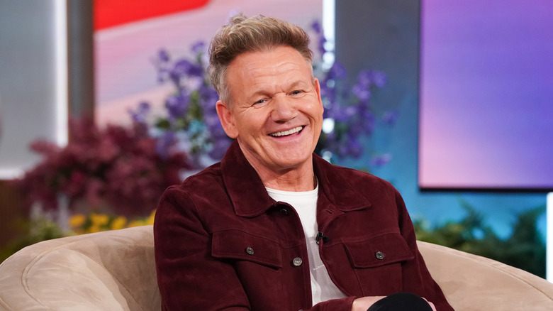 Gordon Ramsay smiles in burgundy jacket with white shirt while sitting on beige chair