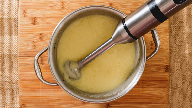 immersion blender in silver pot with yellow soup on wooden chopping board