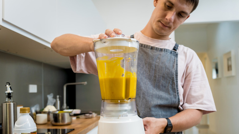 man wearing apron in kitchen using a blender half full with orange mixture
