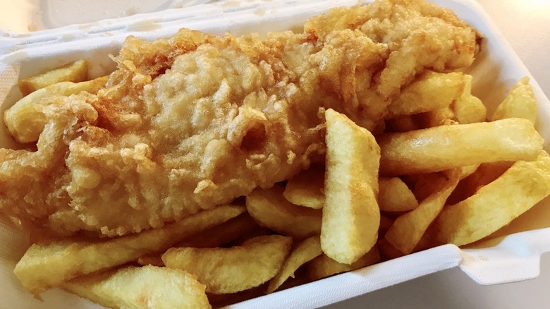 A piece of fried fish sitting in a cardboard container or top some thick cut french fries