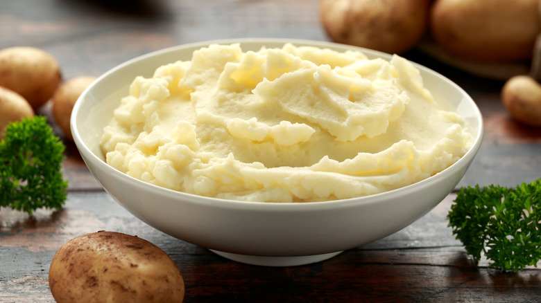 A bowl of mashed potatoes sitting on a wooden table with some whole potatoes and parsley nearby