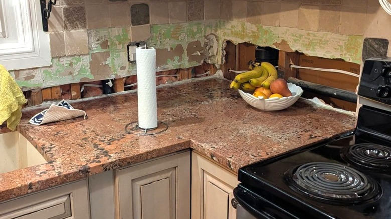Kitchen with salmon pink granite counters holding fruit bowl and paper towel holder, black stove, white cabinets, and distressed blush and green toned backsplash