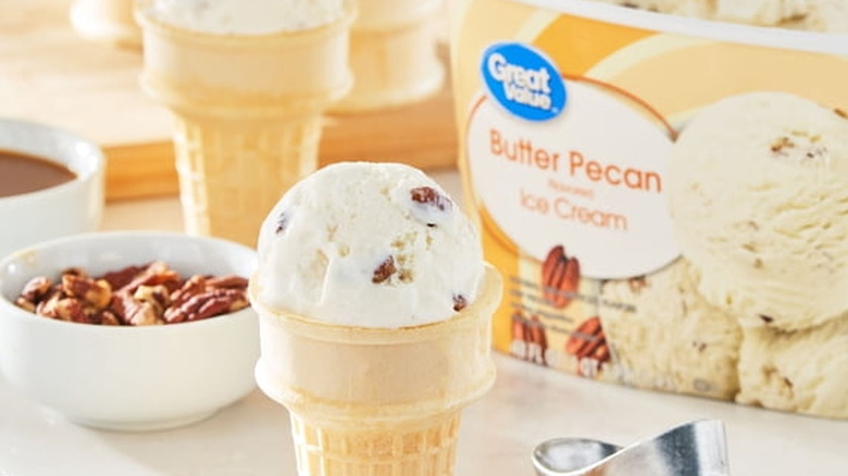 A scoop of Walmart Great Value Butter Pecan Ice Cream on a cone next to a scoop in front of a carton of the ice cream and a bowl of pecans