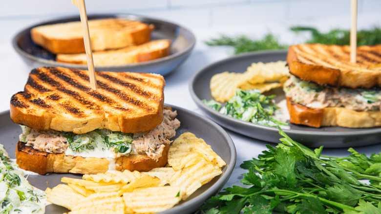 Two grilled salmon salad sandwiches with chips and salad on two plates.