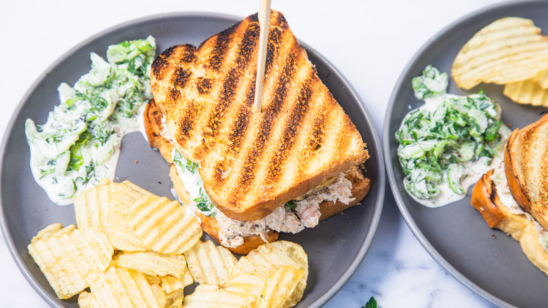 Two grilled salmon salad sandwiches with chips and salad on two plates.