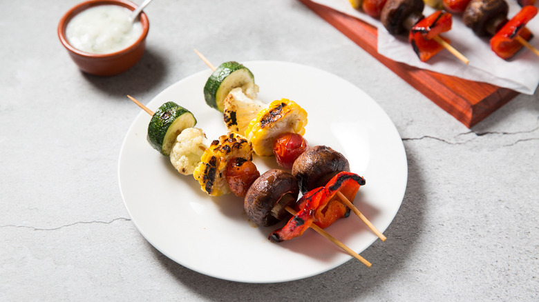 Two veggie skewers served on white plate next to herby yogurt sauce in a small bowl