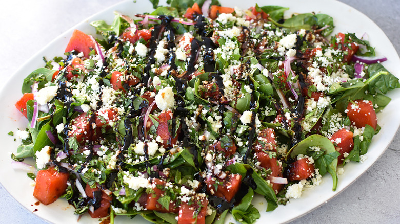 A salad composed of grilled watermelon, cotija cheese, mint, basil, baby spinach, and balsamic vinegar presented on a white plate 