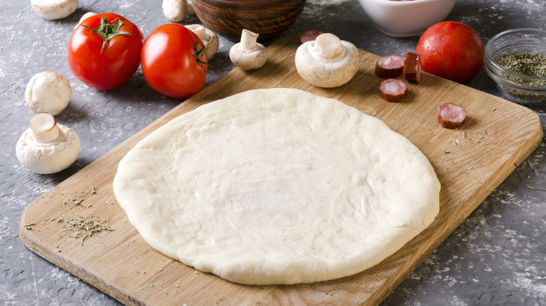 Shaped round of raw pizza dough on wooden board next to mushrooms, tomatoes, and slices of sausage