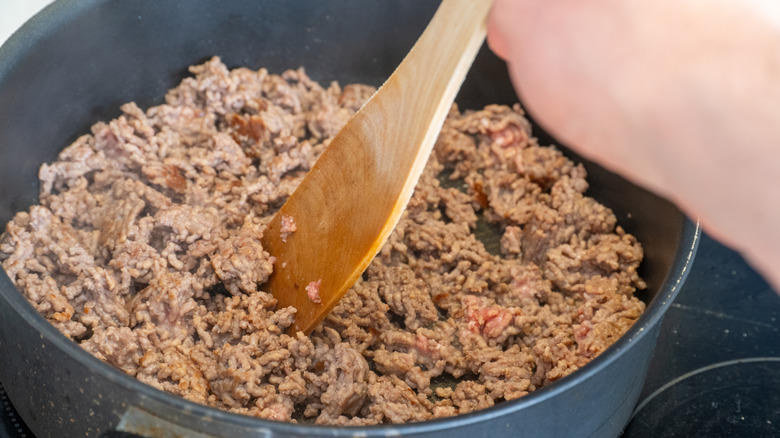 A person using a wooden spoon to stir ground beef browning in a pot