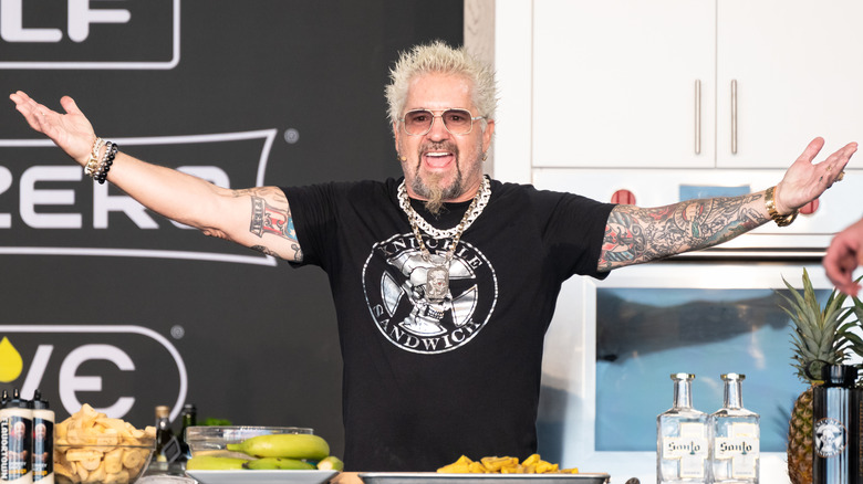 Guy Fieri smiling with arms wide in front of a table with bottles and fruits