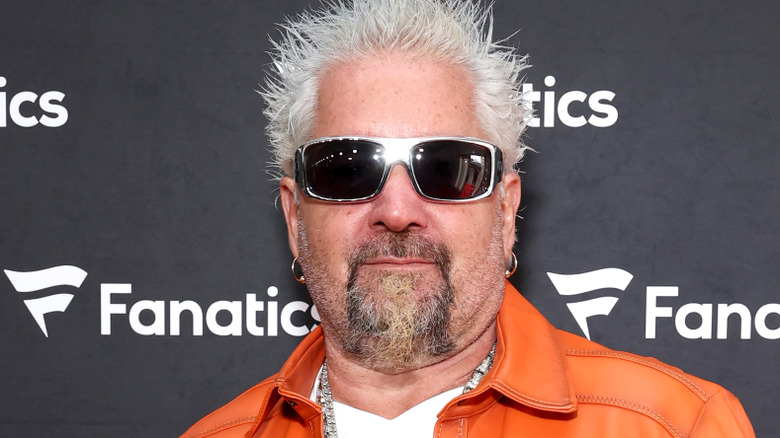 Guy Fieri with orange jacket and sunglasses, smiling behind black background