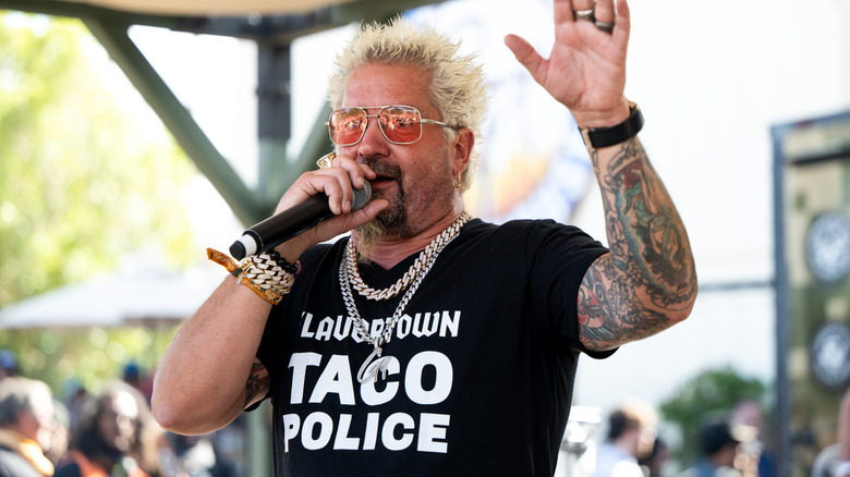 Guy Fieri holds a microphone while wearing a T-shirt reading 
