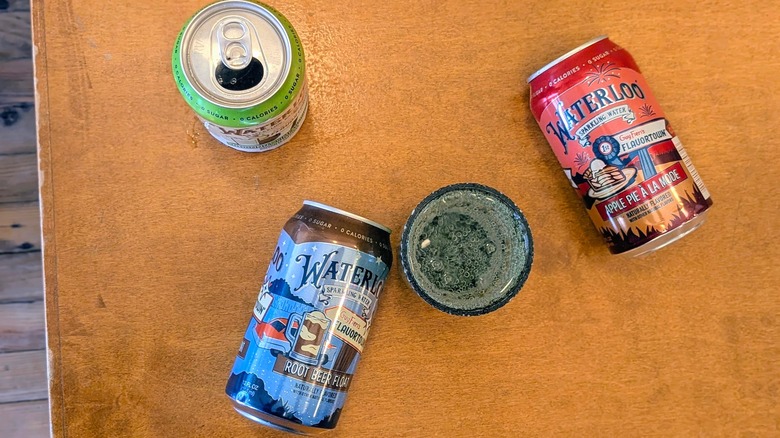 Three cans of Waterloo sparkling water, two overturned, and a plastic cup of sparkling water on a wood table