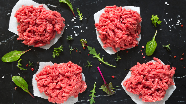Minced beef portioned for four burgers on paper and black background with greens