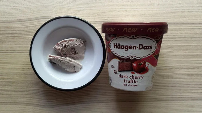 A small bowl of Häagen-Dazs Dark Cherry Truffle ice cream, sitting next to a tub of that ice cream on a wooden surface