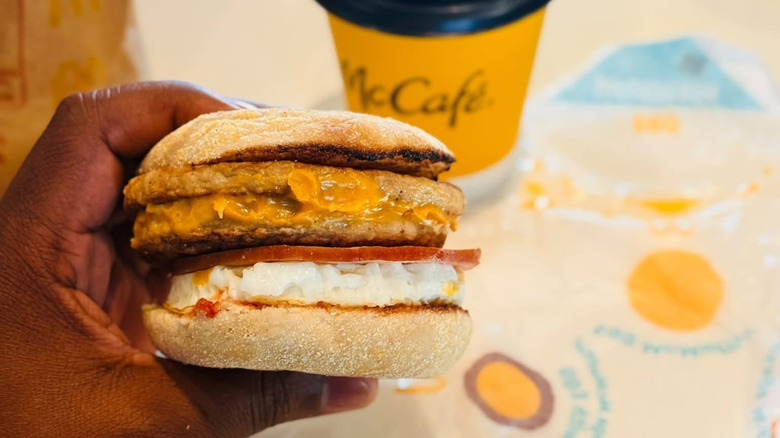 South African Mega McMuffin featuring two sausage patties, chicken roll, egg, cheese, on an English muffin being held in someone's hand with a McCafe coffee in the background