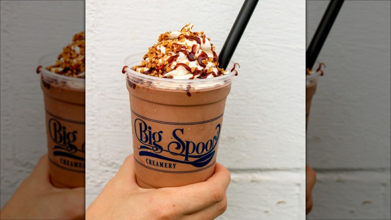 Hand holding milkshake topped with whipped cream, chopped nuts, and chocolate syrup
