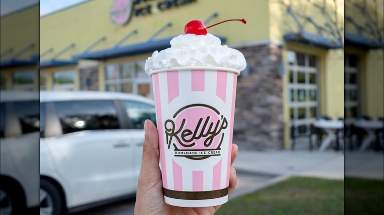 Hand holding milkshake topped with whipped cream and cherry in parking lot