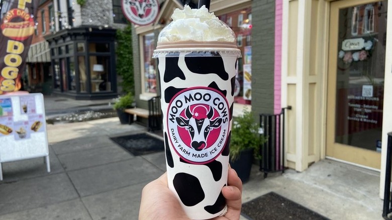 Hand holding chocolate milkshake topped with whipped cream in front of ice cream shop