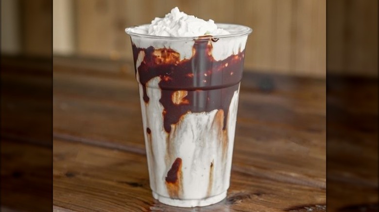 Milkshake topped with whipped cream, hot fudge showing through sides of plastic cup