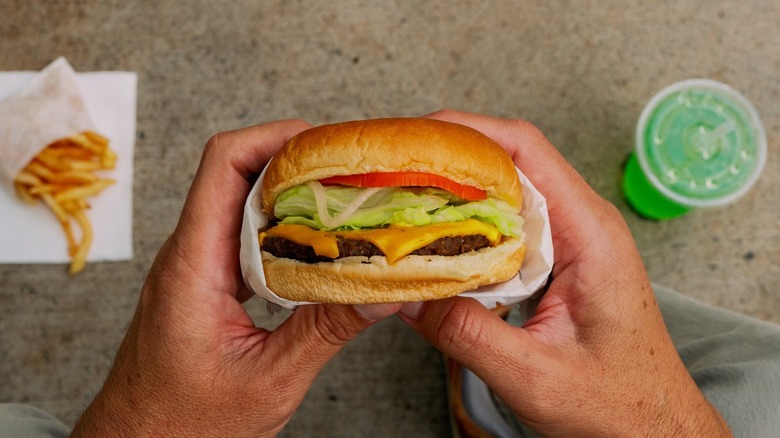 a pair of hands holding a W&M burger with fries and a green drink in the background