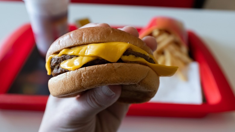 a hand holding cheeseburger in front of a tray with fries and a drink