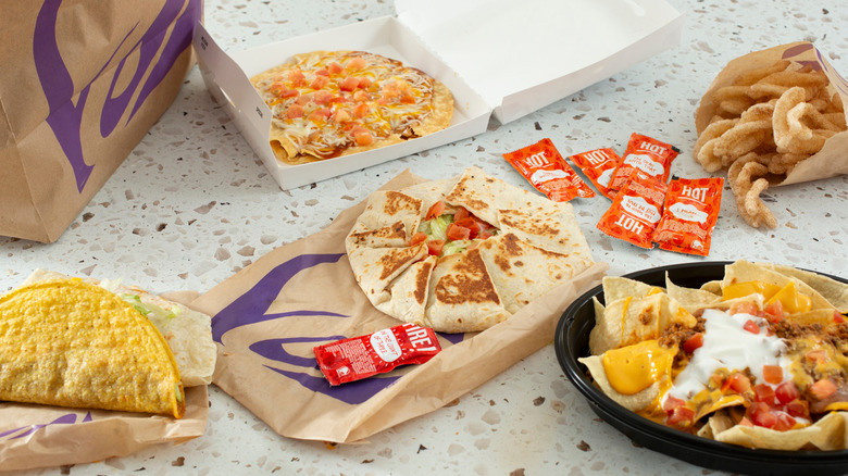 Many Taco Bell items on a table, including a Mexican pizza, hard and soft shell tacos, a crunchwrap, cinnamon twists and nachos