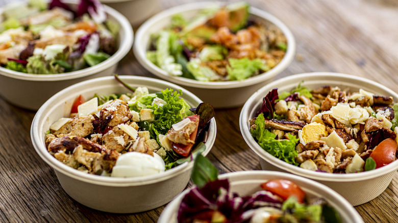 Multiple salads in paper bowls on a wooden table.