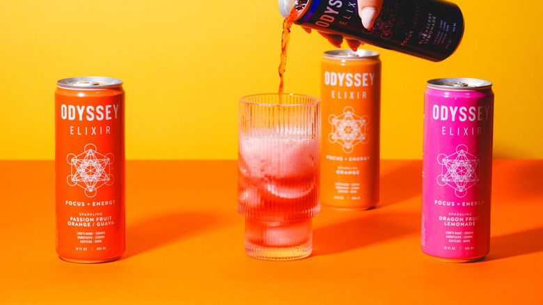 Person pouring a can of Odyssey Elixir into a glass cup surrounded by other Odyssey canned drinks