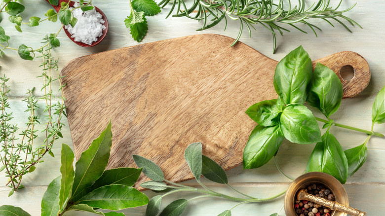 A wooden cutting board surrounded by sprigs of fresh green herbs