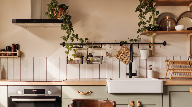 Empty kitchen island with marble surface in foreground, green vintage countertop with drawers and pendant lights hanging above, lots of flowers in jars, blurred background