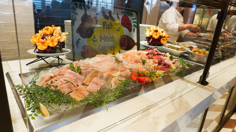 A cruise line buffet featuring raw fish behind glass, garnished with greens, lemon, and small tomatoes, with Edible Arrangements in the background