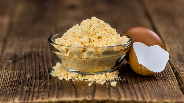 A small glass bowl of fine, yellow powdered egg next to a cracked egg shell. Both are sitting on a wooden table top, with some powdered egg scattered around the bottom of the bowl