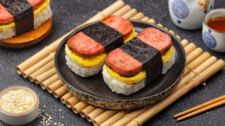 Two pieces of Spam musubi on black plate on top of bamboo mat