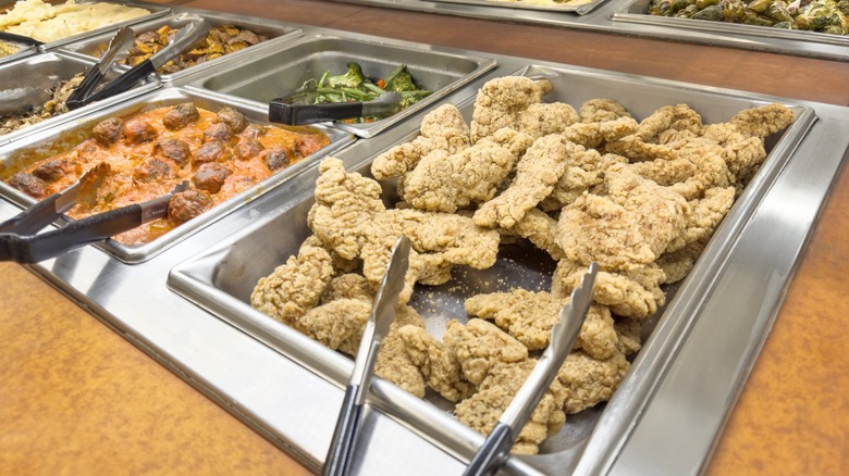 Buffet featuring fried chicken in a warming tray with tongs, also meatballs, broccoli, and other dishes