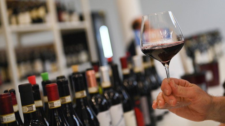Hand holds glass of red wine in front of bottles at shop.