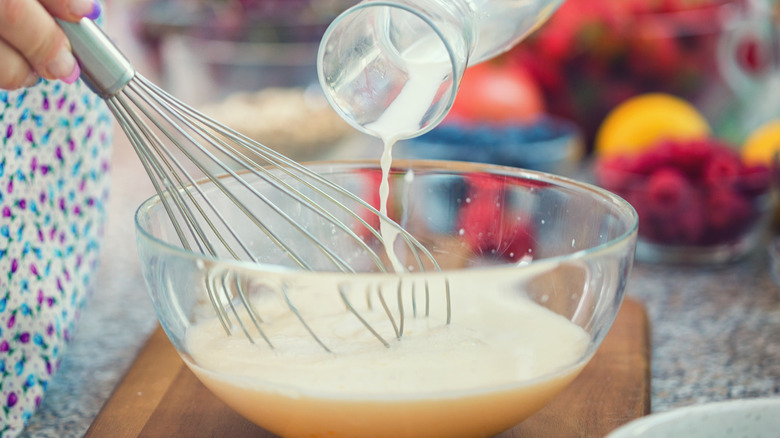 A cook prepares pancake batter, whisking a mixing bowl with one hand while pouring milk with the other
