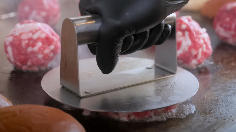 A gloved hand uses a burger press to smash ground beef balls on a griddle