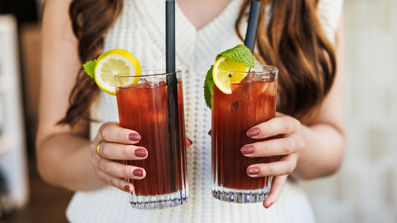 Woman holding iced tea glasses with lemon garnishes and black straws.