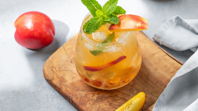 Iced tea with peaches and mint in a glass, with a wooden cutting board, grey linen towel and a whole peach nearby.