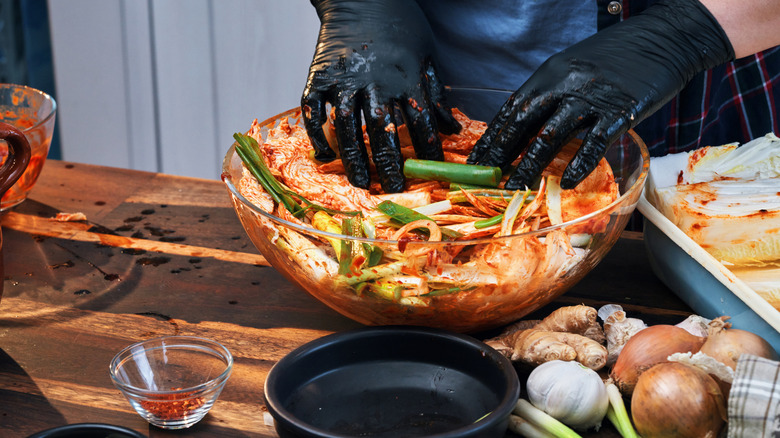 The person wearing gloves making homemade kimchi in a large glass bowl. There are spices, cabbage, onions, garlic, and ginger on the table nearby