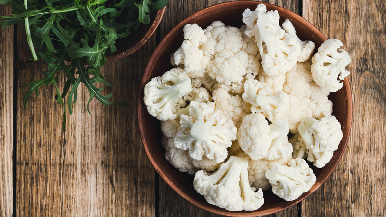 Cauliflower florets in a bowl, next to arugula, on rustic wooden background