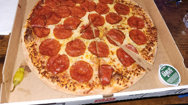 A Papa Johns pepperoni pizza sitting in the box with a tub of garlic sauce and a yellow pepperoncini pepper