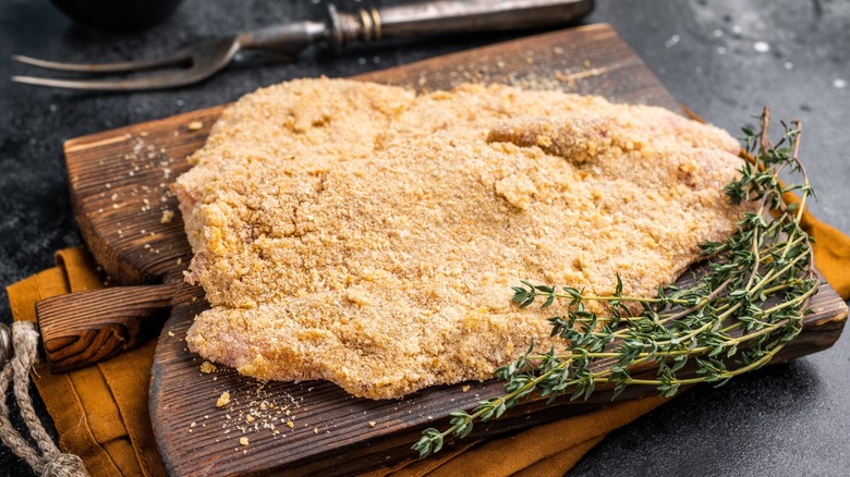 A breaded pork schnitzel on a wooden cutting board with some tarragon