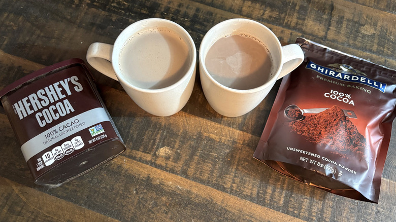 Hershey's and Ghirardelli cocoa powder containers with hot chocolates in white mugs on wooden table