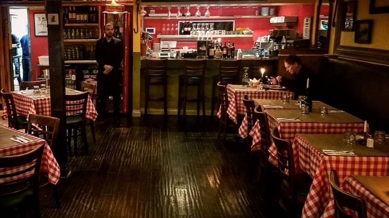 a plate of spaghetti bolognese from La Pergoletta on a red checkered tablecloth with wine bottles, desserts, etc. behind it