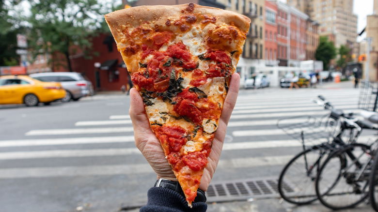 Hand holding a slice of pizza up in front of a busy NYC street